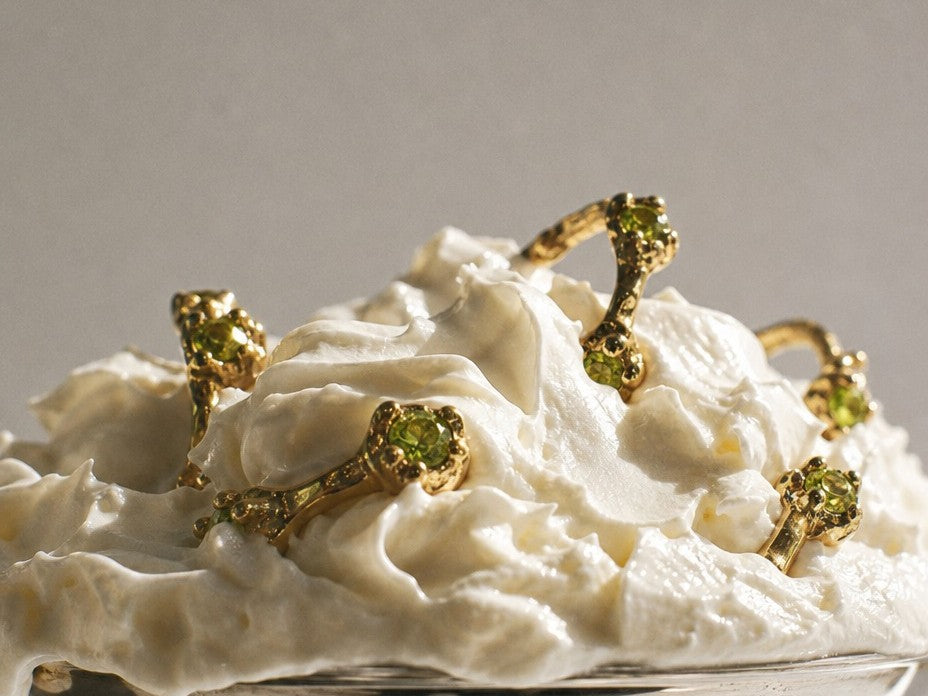 Several gold rings with green gemstones are arranged on top of a mound of whipped cream against a plain light gray background.